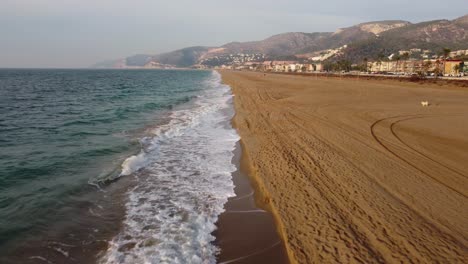 Castelldefels-Beach-in-Barcelona-with-waves-washing-ashore,-empty-sandy-beach,-coastal-town-backdrop,-aerial-view