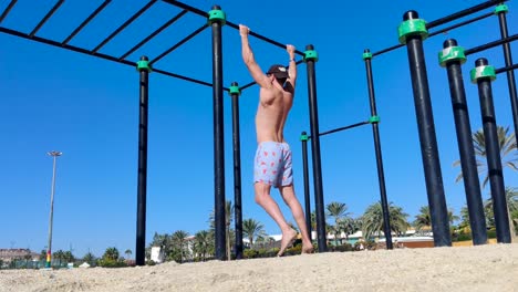 A-young-male-wearing-shorts,-a-baseball-hat-and-sunglasses-doing-pull-ups-on-bars-at-an-outdoor-gym-on-the-beach