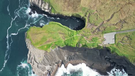 Drone-shot-of-the-Kilkee-Cliffs-of-Ireland,-where-the-ocean-meets-the-countryside-fields-and-waves-are-crashing-against-the-tall-rugged-coastline