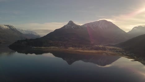 Panning-drone-shot-showing-Beautiful-mountain-range-of-Scotland-with-snowy-mountaintop-at-sunny-day---Lake-reflection-of-mountains-on-water-surface