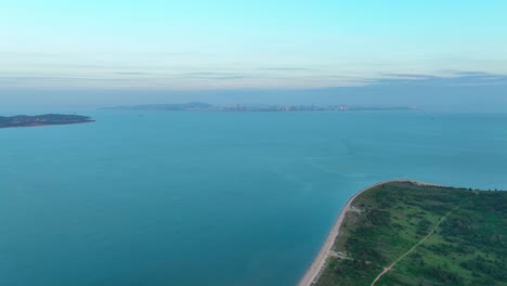 Rückwärtsaufnahme-Aus-Der-Luft,-Die-Den-Sandstrand-Auf-Der-Grünen-Insel-Kinmen-金門-Quemoy-Und-Die-Stadt-Xiamen-Im-Hintergrund-Mit-Wunderschönem-Meer-An-Einem-Nebligen-Tag-Zeigt