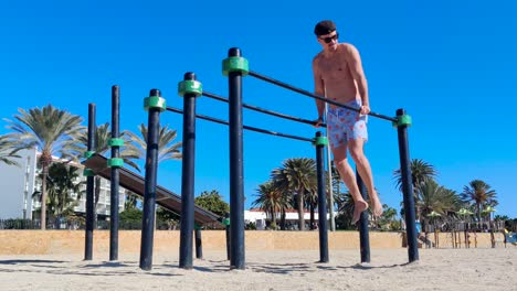 A-young-male-wearing-shorts,-a-baseball-hat-and-sunglasses-doing-triceps-dips-on-parallel-bars-at-an-outdoor-gym-on-the-beach