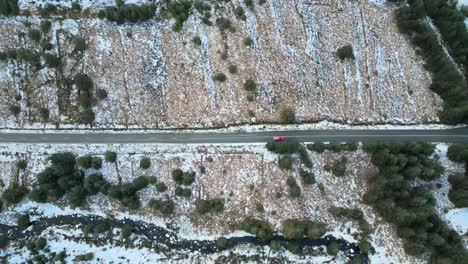 Solitary-red-car-driving-on-a-snow-dusted-road-flanked-by-winter-trees,-aerial-view