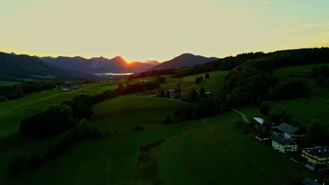 Aerial-drone-backward-moving-shot-over-green-farmlands-along-mountain-slope-in-Norway-with-sun-rising-in-the-background