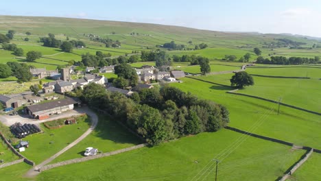 Drone-footage-reversing-and-panning-showing-rural-Yorkshire-countryside,-revealing-a-small-country-village-and-campsite,-including-farmland,-dry-stone-walls-and-Ingleborough-mountain-in-the-distance