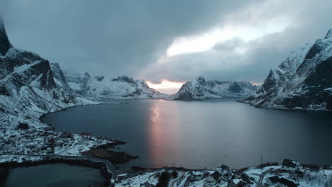 Drone-flight-over-Reine,-Lofoten-Islands,-Norway-during-sunset-with-the-sun-peeking-through-dark-clouds,-snowy-mountains,-and-a-moody-ocean-scene