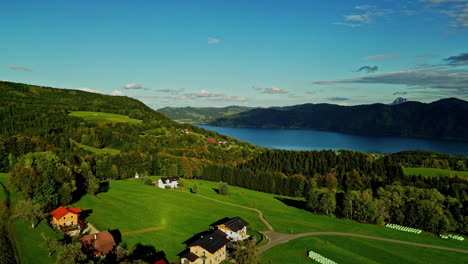 Lush-countryside-with-green-fields,-a-tranquil-lake,-and-forested-hills-under-a-clear-sky,-aerial-view