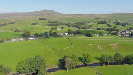 Drone-footage,-moving-and-panning-showing-the-mountain-of-Ingleborough-in-the-distance-with-a-rural-Yorkshire-countryside-village-and-river-on-a-sunny-summer-day