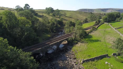 Aerial-footage-reversing-over-a-river-with-exposed-river-bed-in-a-shallow-valley-in-rural-Yorkshire-UK-with-railway-bridge-and-tracks-in-front-and-flying-just-over-a-tree-with-mountains-in-distance