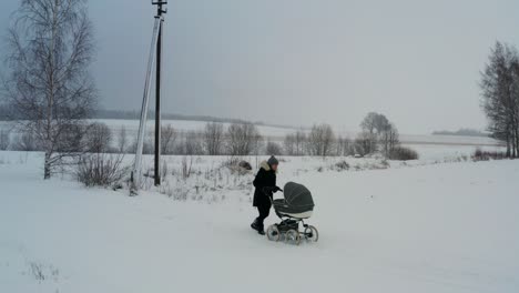 Female-in-black-jacket-walk-with-baby-stroller-on-snowy-road,-winter-season