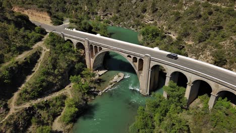 Murillo-de-Gallego-Bridge-in-Huesca-over-turquoise-river,-surrounded-by-lush-greenery,-aerial-view
