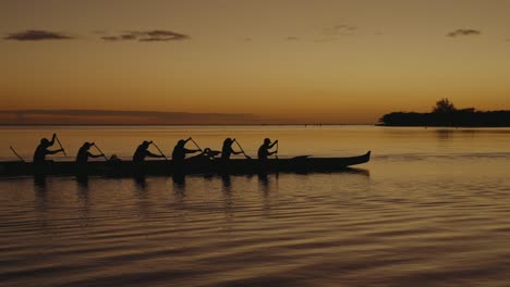 a-6-person-team-paddles-an-outrigger-canoe-or-wa'a-across-a-calm-ocean-in-the-Hawaiian-islands-at-sunset-as-other-boats-follow-behind-silhouette
