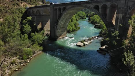 Murillo-de-Gallego-Bridge-in-Huesca-over-a-turquoise-river-surrounded-by-greenery,-aerial-view