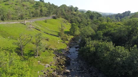 Drone-footage-on-a-sunny-summer-day-through-a-shallow-river-valley-and-exposed-river-bed,-lined-with-grassy-banks-and-trees-with-a-country-road-on-one-side-and-railway-on-the-other