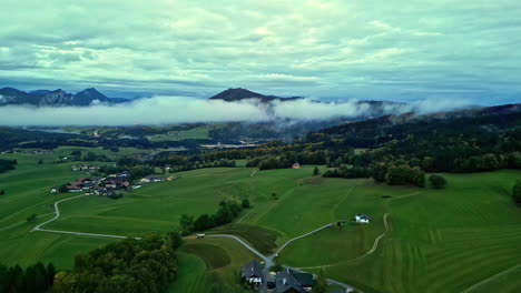 Neblige-Landschaft-Im-Morgengrauen-Mit-Grünen-Feldern,-Verstreuten-Häusern-Und-Hügeln-Im-Hintergrund