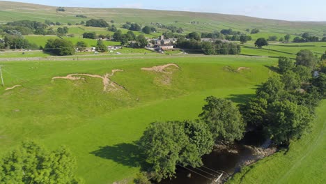 Drone-footage-flying-parallel-to-a-tree-lined-river-in-rural-Yorkshire-near-Ingleton-on-a-sunny-summer-day,-surrounded-by-fields,-countryside-and-a-small-village-with-hills-in-the-background