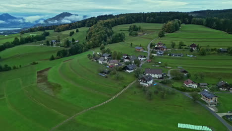 Ruhiges-Ländliches-Dorf-Mit-Grünen-Feldern,-Häusern-Und-Kurvenreichen-Straßen-Vor-Der-Kulisse-Ferner-Berge
