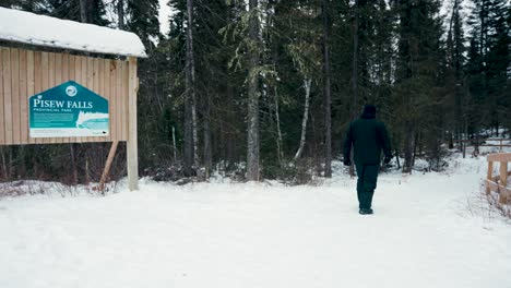 The-Camera-Follows-an-Aboriginal-Indigenous-Man-Wearing-Black-Winter-Clothes-Walks-Past-the-Entry-Sign-for-Pisew-Falls-Provincial-Park-Near-Thompson-Northern-Manitoba-Canada-To-Hike-down-the-Trail