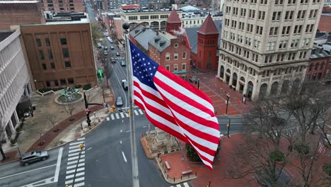 Aerial-establishing-shot-of-American-flag-waving-over-city-square-during-winter