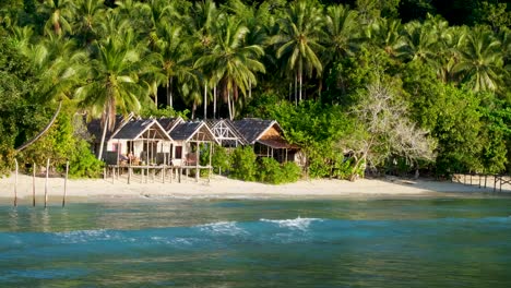 Simple-wooden-huts-with-rainforest-trees-on-white-sandy-beach-overlooking-ocean-water-on-remote-tropical-island-in-Raja-Ampat,-West-Papua,-Indonesia