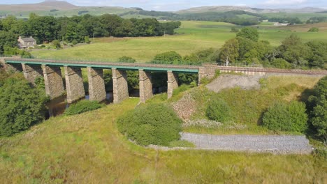 Drone-footage-on-a-sunny-day-moving-sideways-and-panning-parallel-to-a-rural-railway-line-and-stone-railway-bridge-in-the-Yorkshire-countryside-near-Settle