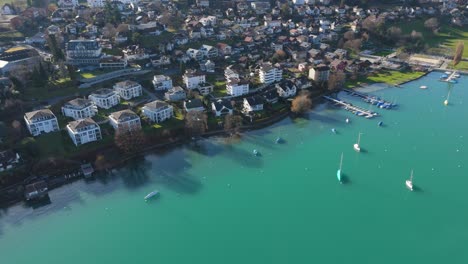 Spiez-town-by-Lake-Thun-with-Swiss-Alps-backdrop,-tranquil-boats-and-clear-waters,-sunny-day,-aerial-view