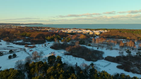 Aerial-approaching-shot-of-colorful-trees-in-winter-landscape-with-przymorze-district-of-Gdansk-city---Baltic-Sea-and-Horizon-at-sunset-time-in-background