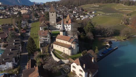 Spiez-castle-and-church-with-Lake-Thun-and-Swiss-Alps-in-the-background,-clear-day,-aerial-view