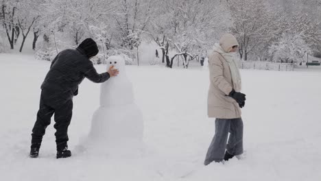 Couple-Making-Snowman-In-Public-Park-On-A-Snowy-Day