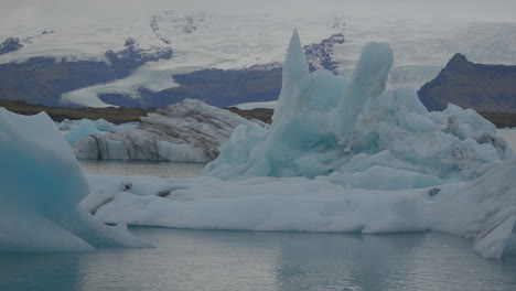 Glaciares-Flotando-En-Una-Laguna-Glaciar,-Islandia,-Con-Gaviotas-Volando-Sobre-Sus-Cabezas,-Con-El-Telón-De-Fondo-De-Aguas-Azules-Que-Se-Mueven-Hacia-La-Playa-De-Diamantes.