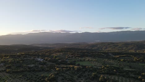 Luftflug-Mit-Einer-Drohne-In-Einem-Tal-In-Spanien-über-Einem-Wald-Einheimischer-Bäume-Des-Landes,-Der-Bei-Sonnenuntergang-Sanft-Nach-Links-Abbiegt,-Mit-Einem-Wunderschönen-Hintergrund-Aus-Bergen