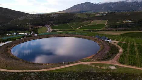 Perfectly-round,-man-made-dam-on-a-wine-farm-reflects-the-cloudy-sky-like-a-mirror,-in-between-the-vineyards-with-mountains-in-the-distance