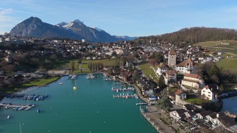 Spiez-town-with-Lake-Thun-and-Swiss-Alps-in-background,-clear-sky,-boats-on-water,-aerial-view