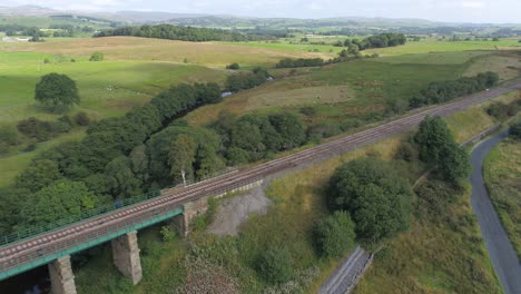 Drone-footage-moving-parallel-to-a-rural-railway-line-over-a-stone-bridge-and-river-with-fields,-farmland,-hills-and-countryside-all-around