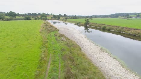 Drone-footage-flying-low-and-following-a-shallow-slow-moving-river-in-the-Lake-District-countryside,-UK-showing-the-exposed-river-ban,-fields,-hills-and-cattle-in-the-distance