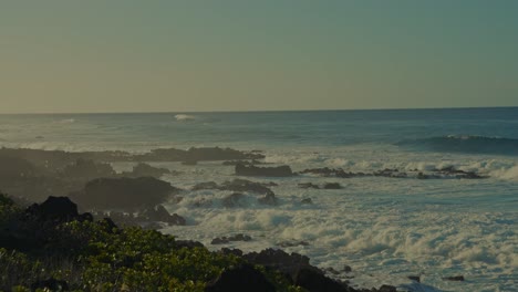 the-white-caps-on-the-blue-ocean-waves-gather-at-the-shore-along-the-island-coast-of-Kaena-point-on-Oahu,-Hawaii-as-the-sun-begins-to-set