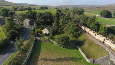 Beautiful-summer-evening-drone-footage-elevating-and-slowly-panning-at-the-village-of-Selside,-Yorkshire,-UK,-fields,-farmland-and-rolling-hills-as-a-freight-train-passes-under-and-into-the-distance