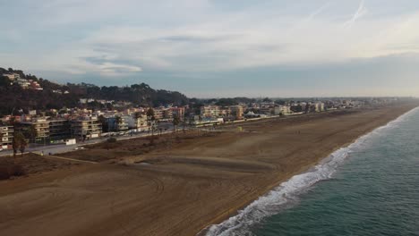 Castelldefels-Beach-in-Barcelona-with-waves-lapping-the-shore,-buildings-in-the-background,-clear-sky,-aerial-view