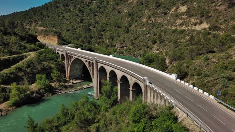 Murillo-de-Gallego-Bridge-in-Huesca-with-cars-passing-over-and-turquoise-river-flowing-underneath,-sunny-day,-aerial-view