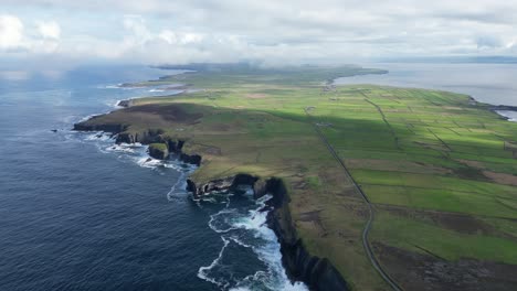 Loophead-peninsula-with-dramatic-cliffs,-lush-green-fields,-and-coastal-roads-under-a-cloudy-sky,-aerial-view