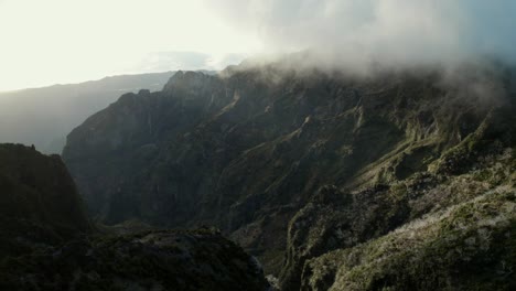 Drone-flying-between-rugged-mountains-during-sunrise-at-Madeira,-while-fog-and-clouds-comes-into-the-picture