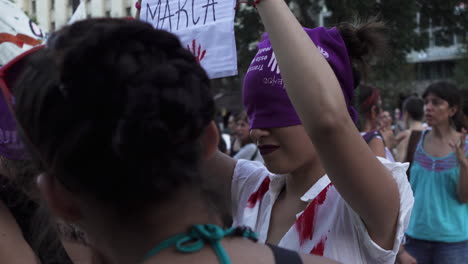 Young-woman-performs-public-manifestation,-holds-poster-with-her-eyes-covered-by-purple-feminist-bandana,-she-has-red-stains-representing-blood-and-violence