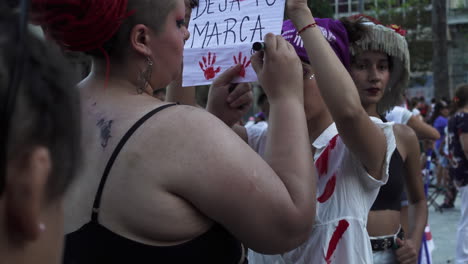 Dos-Mujeres-Manifestantes-Con-Letrero,-Tienen-Manchas-Rojas-Que-Imitan-Sangre,-Realizan-Manifestación-Pública-De-Descontento.