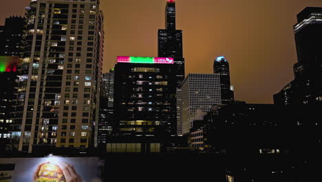 Aerial-view-rising-in-front-of-the-Willis-Tower-from-West-Loop-Gate,-cloudy-night-in-Chicago