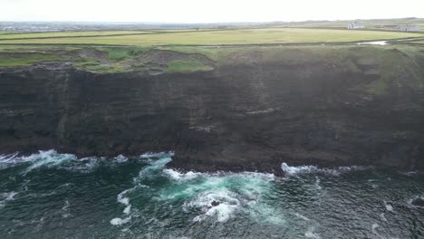 Kilkee-Cliffs-with-waves-breaking,-lush-green-fields-in-the-backdrop-from-drone-perspective