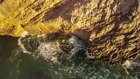 Waves-crashing-cliffs-on-sunny-evening,-aerial-downwards-view