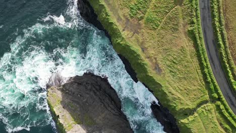 Waves-crashing-against-rocky-cliffs-with-lush-green-landscape-at-Kilkee-cliffs,-aerial-view