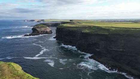 Aerial-view-of-rugged-cliffs-by-the-sea-with-waves-crashing,-green-countryside-in-the-background
