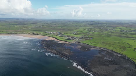 Doughmore-Bay-with-waves-hitting-the-shore,-green-countryside-backdrop,-and-cloudy-skies,-aerial-view