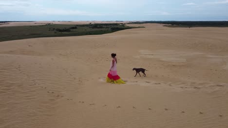 Woman-in-long-flowing-dress-walks-on-massive-sand-dune-with-her-dog